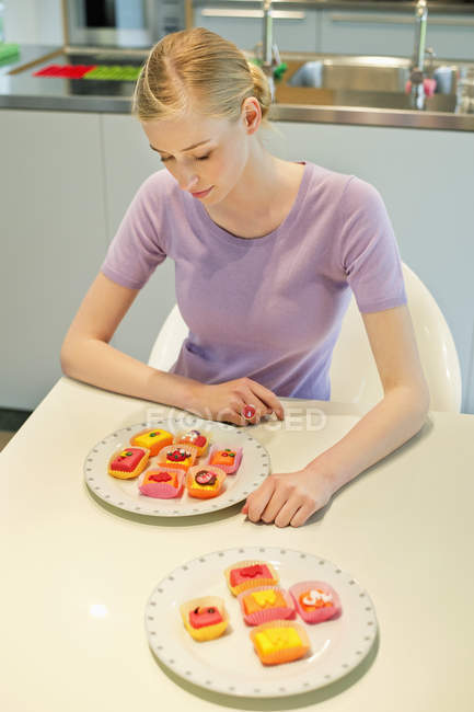 Young blond woman sitting at kitchen table with colorful cupcakes — Stock Photo