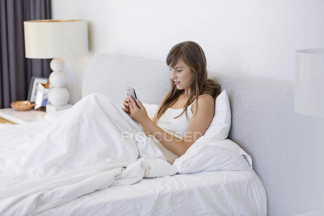 Jovem mulher sentada na cama sob cobertor e usando telefone celular — Fotografia de Stock