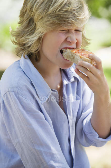 Мальчик с светлыми волосами ест сэндвич на открытом воздухе — стоковое фото