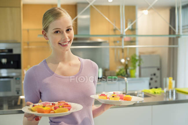 Happy woman holding two plates of homemade cupcakes in kitchen — Stock Photo