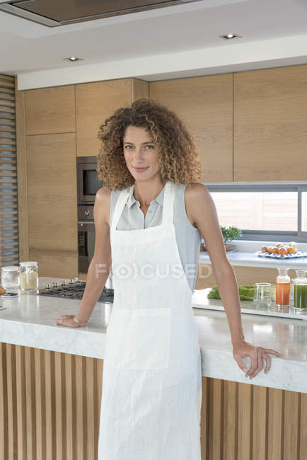 Портрет улыбающейся женщины в фартуке, прислонившейся к кухонной стойке — стоковое фото