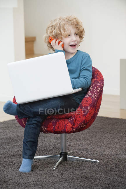 Хлопчик розмовляє на мобільному телефоні і використовує ноутбук у кріслі вдома — стокове фото