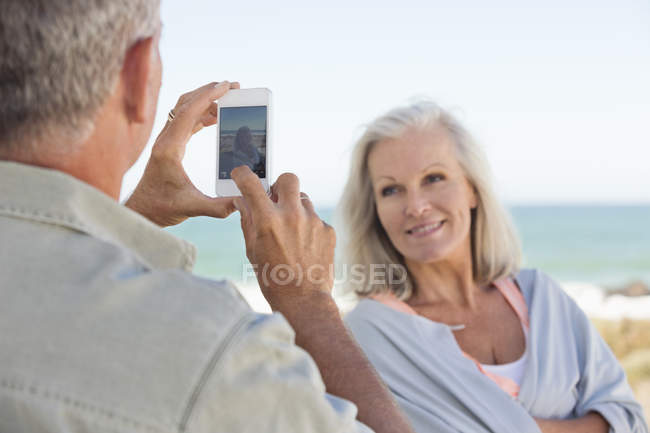 Man taking picture of wife with cell phone on beach — Stock Photo