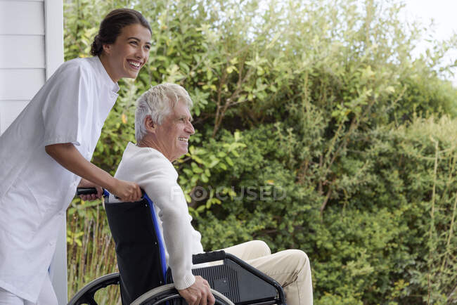 Женщина-медсестра помогает пожилому мужчине в инвалидном кресле на крыльце — стоковое фото