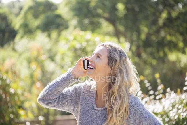 Happy woman in sweater talking on phone in sunny garden — Stock Photo