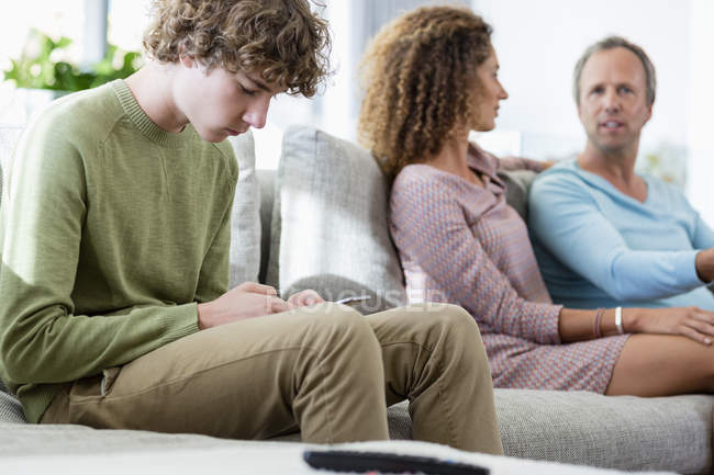 Niño usando el teléfono móvil mientras los padres hablan en segundo plano en la sala de estar en casa - foto de stock
