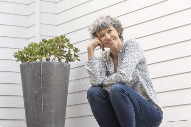 Happy mature woman sitting next to potted plant in front of house wall — Stock Photo