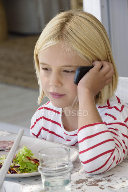 Little girl talking on mobile phone while eating meal — Stock Photo