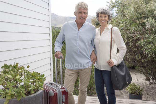 Glückliches Seniorenpaar steht mit Koffer vor dem Haus und blickt in die Kamera — Stockfoto
