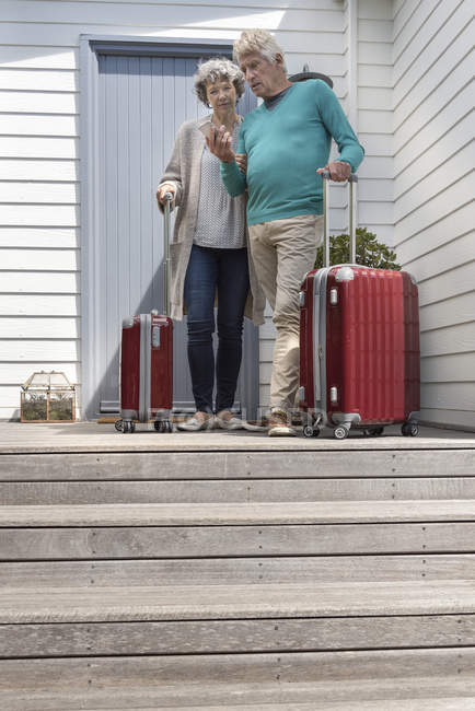 Старшая пара с чемоданами с помощью смартфона у входной двери — стоковое фото