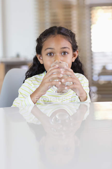 Маленькая девочка пьет воду из стекла за столом дома и смотрит в камеру — стоковое фото