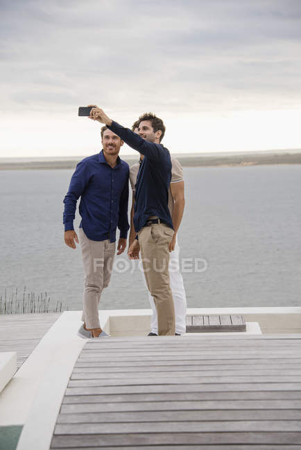 Amigos tomando selfie com telefone celular no terraço de madeira no lago — Fotografia de Stock