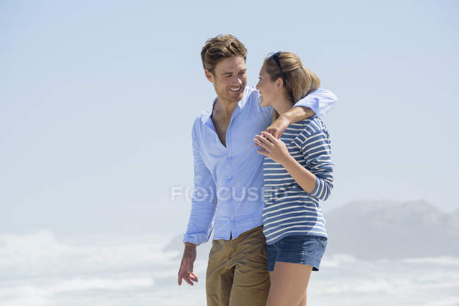 Sonriente pareja romántica caminando en la playa bajo el cielo azul - foto de stock