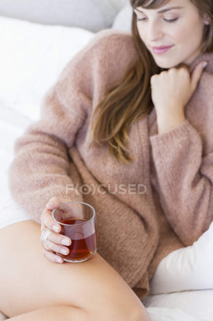 Sonriente joven con taza de té de hierbas sentado en la cama - foto de stock