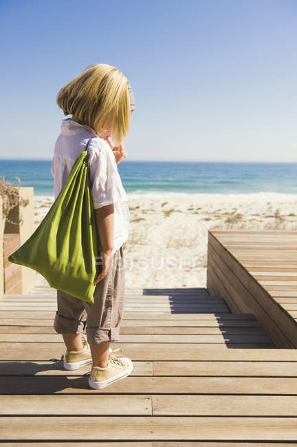 Little blonde girl with bag standing on boardwalk on sandy beach — Stock Photo