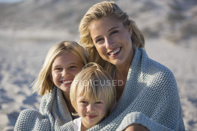 Retrato de uma mãe sorridente e crianças envolto em xale sentado na praia — Fotografia de Stock