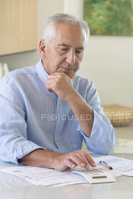 Uomo anziano che usa la calcolatrice mentre fa scartoffie a casa — Foto stock