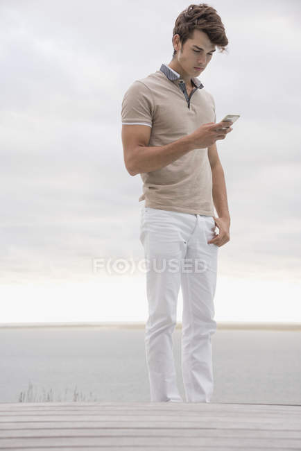 Young man using smartphone on shore under cloudy sky — Stock Photo