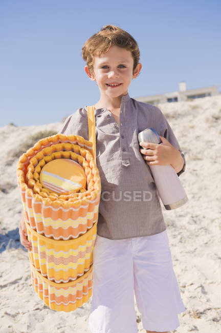 Portrait of smiling little boy carrying mat on beach — Stock Photo