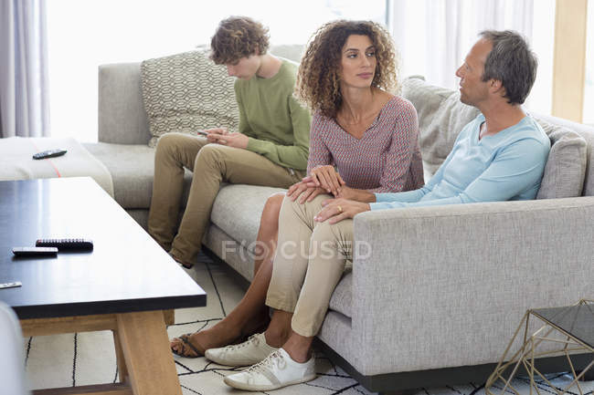 Couple sitting on sofa and talking with son using mobile phone on background in living room at home — Stock Photo