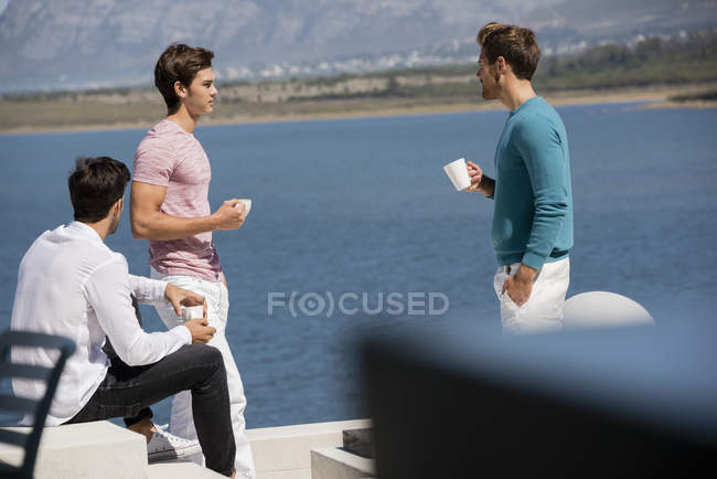 Счастливые друзья-мужчины разговаривают с чашкой кофе на озере — стоковое фото