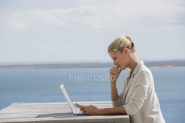 Happy young woman using smartphone at wooden table with laptop on lake shore — Stock Photo
