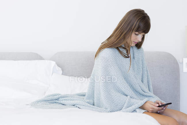 Young woman sitting on bed and using mobile phone — Stock Photo