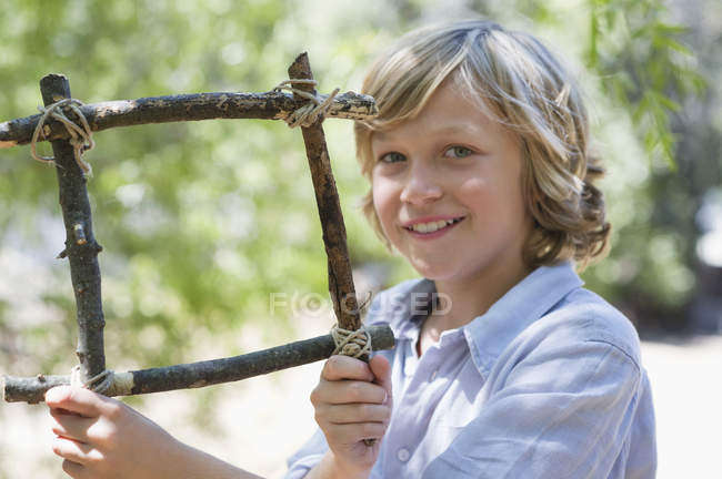 Portrait de garçon blond souriant tenant le cadre de bois flotté à l'extérieur — Photo de stock