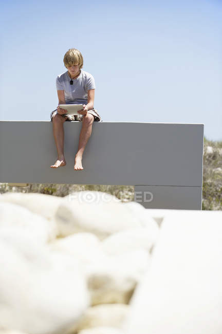Adolescent garçon à l'aide numérique tablette tandis que assis sur mur à l'extérieur — Photo de stock