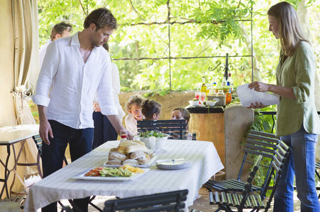 Casal organizando comida na mesa de jantar com a família no fundo — Fotografia de Stock