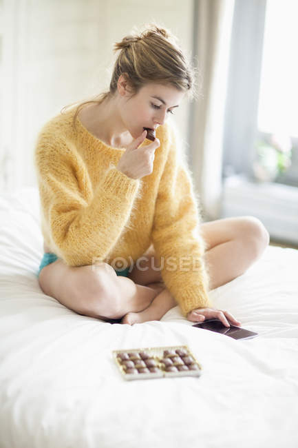 Donna in maglione giallo soffice utilizzando il telefono e mangiare cioccolato sul letto — Foto stock