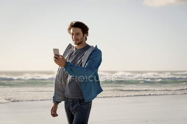 Счастливый молодой человек, использующий телефон с наушниками на пляже — стоковое фото