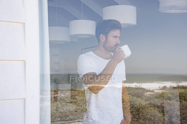 Молодой человек смотрит в окно с чашкой кофе — стоковое фото