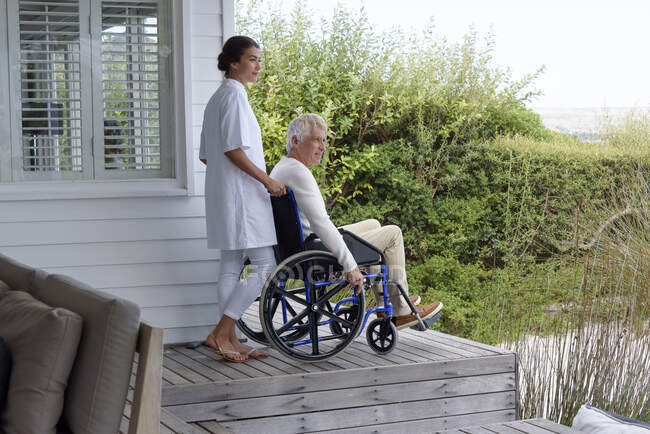 Жіноча медсестра допомагає старшому чоловікові в інвалідному візку на ганку — стокове фото
