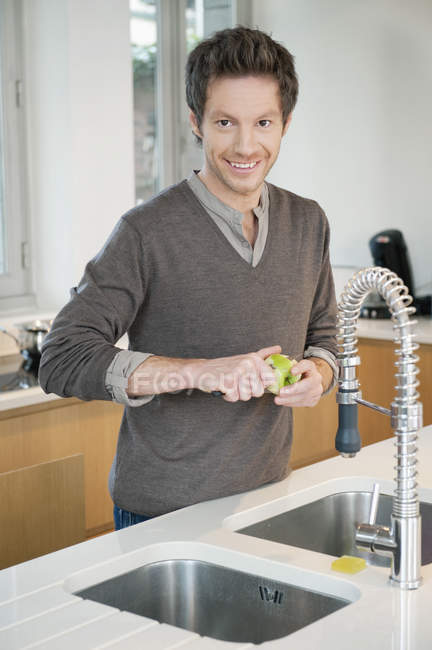 Retrato del hombre sonriente pelando manzana en la cocina - foto de stock