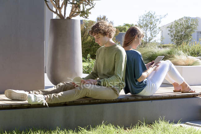 Hermano y hermana sentados espalda con espalda con libro y tableta en el jardín - foto de stock