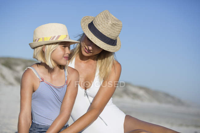 Blond young woman and daughter in hats sitting on beach — Stock Photo