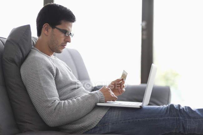 Homem segurando cartão de crédito e usando um laptop no sofá — Fotografia de Stock