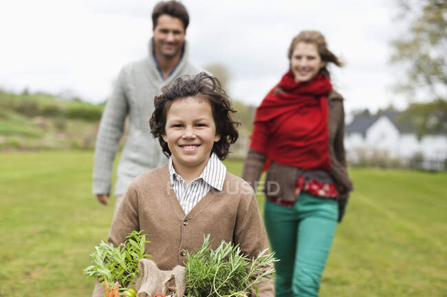 Retrato de un niño sosteniendo una cesta de verduras con sus padres en una granja - foto de stock