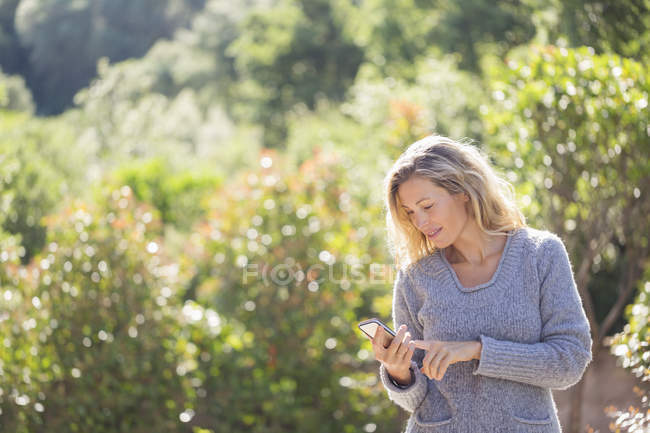 Donna in maglione utilizzando il telefono in giardino soleggiato — Foto stock