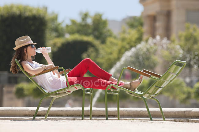 Женщина в соломенной шляпе отдыхает в кресле возле пруда и пьет воду — стоковое фото