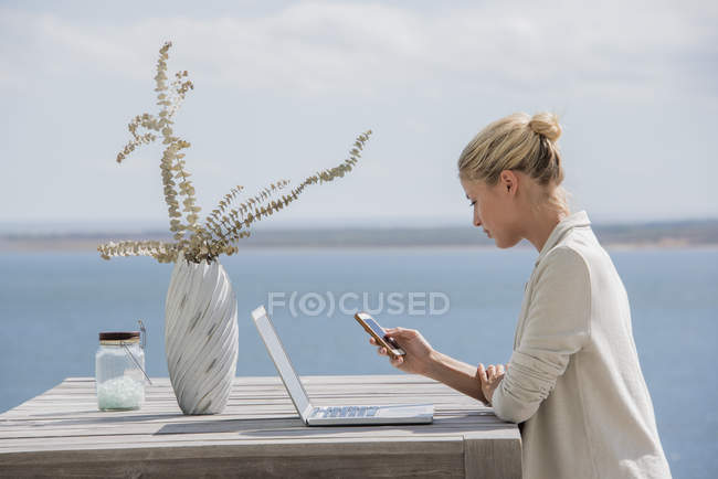 Молодая женщина с помощью смартфона за деревянным столом с ноутбуком на берегу озера — стоковое фото