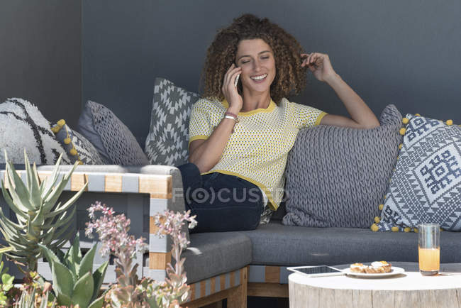 Mulher sorrindo falando no telefone celular enquanto sentado no sofá em casa — Fotografia de Stock