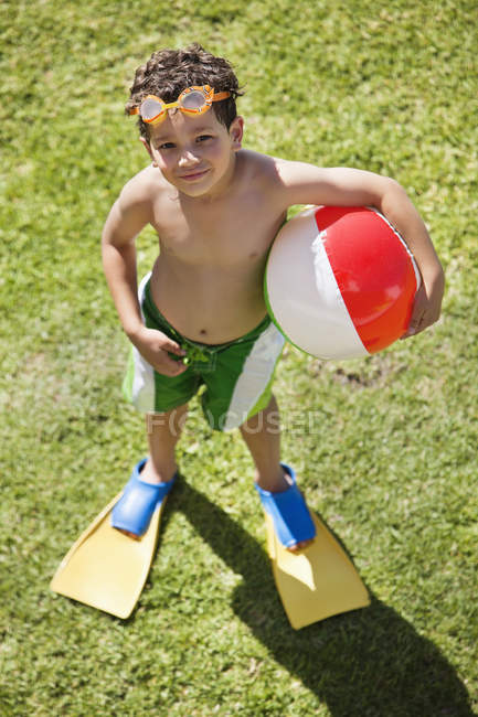 Маленький мальчик в ластах и плавательных очках, держащий пляжный мяч на зеленой лужайке — стоковое фото