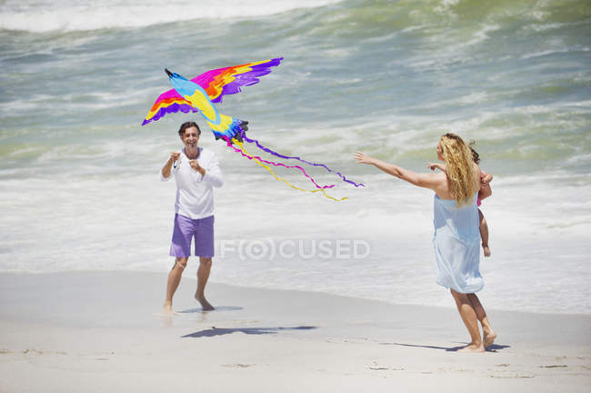 Mother carrying child while man flying kite on beach — Stock Photo