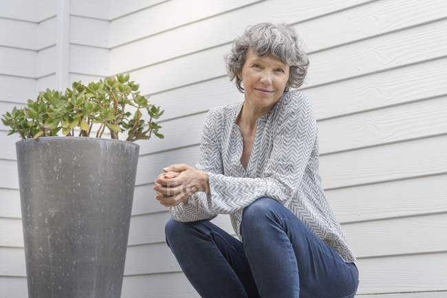 Happy mature woman sitting next to potted plant in front of house wall — Stock Photo