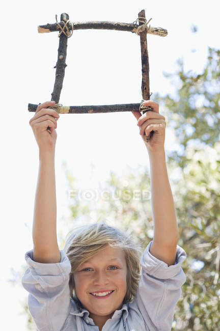 Portrait of cute boy holding frame of driftwood with arms raised outdoors — Stock Photo
