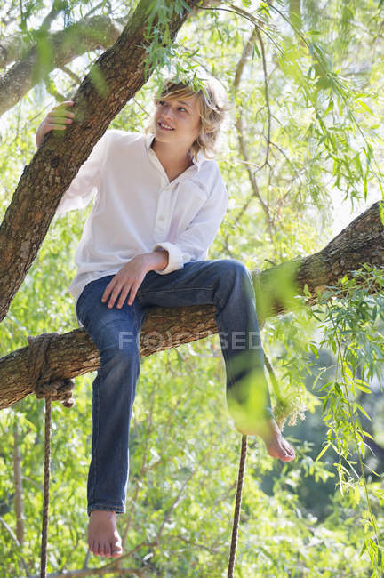 Contemplative teenage boy sitting on tree branch in summer — Stock Photo
