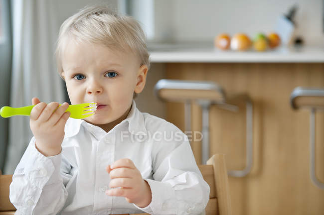 Retrato de un niño rubio comiendo con tenedor en casa - foto de stock