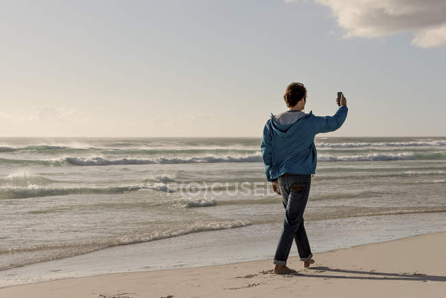 Hombre joven usando el teléfono móvil mientras camina en la playa - foto de stock